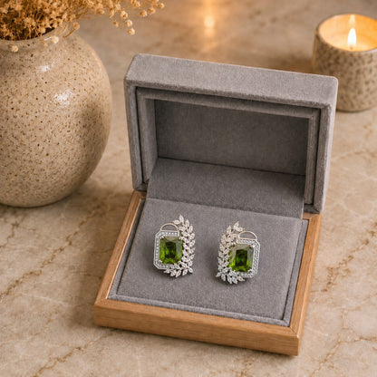 A pair of silver-toned stud earrings with green rectangular gemstones and leaf-shaped silver accents, displayed in an open gray jewelry box on a marble surface with a lit candle and vase in the background.
