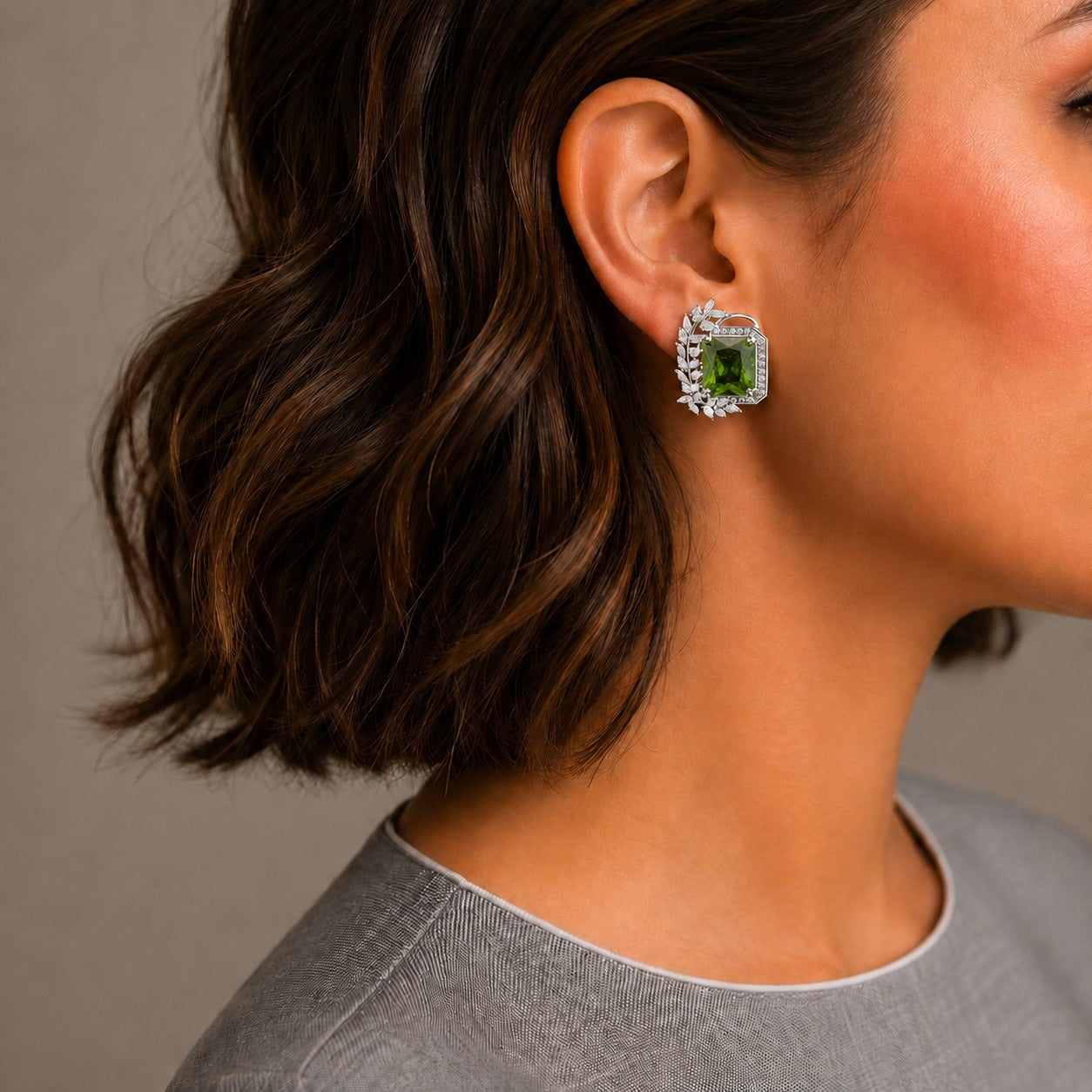 A close-up side profile of a woman with short brown hair wearing a silver-toned stud earring with a green rectangular gemstone surrounded by small clear stones.