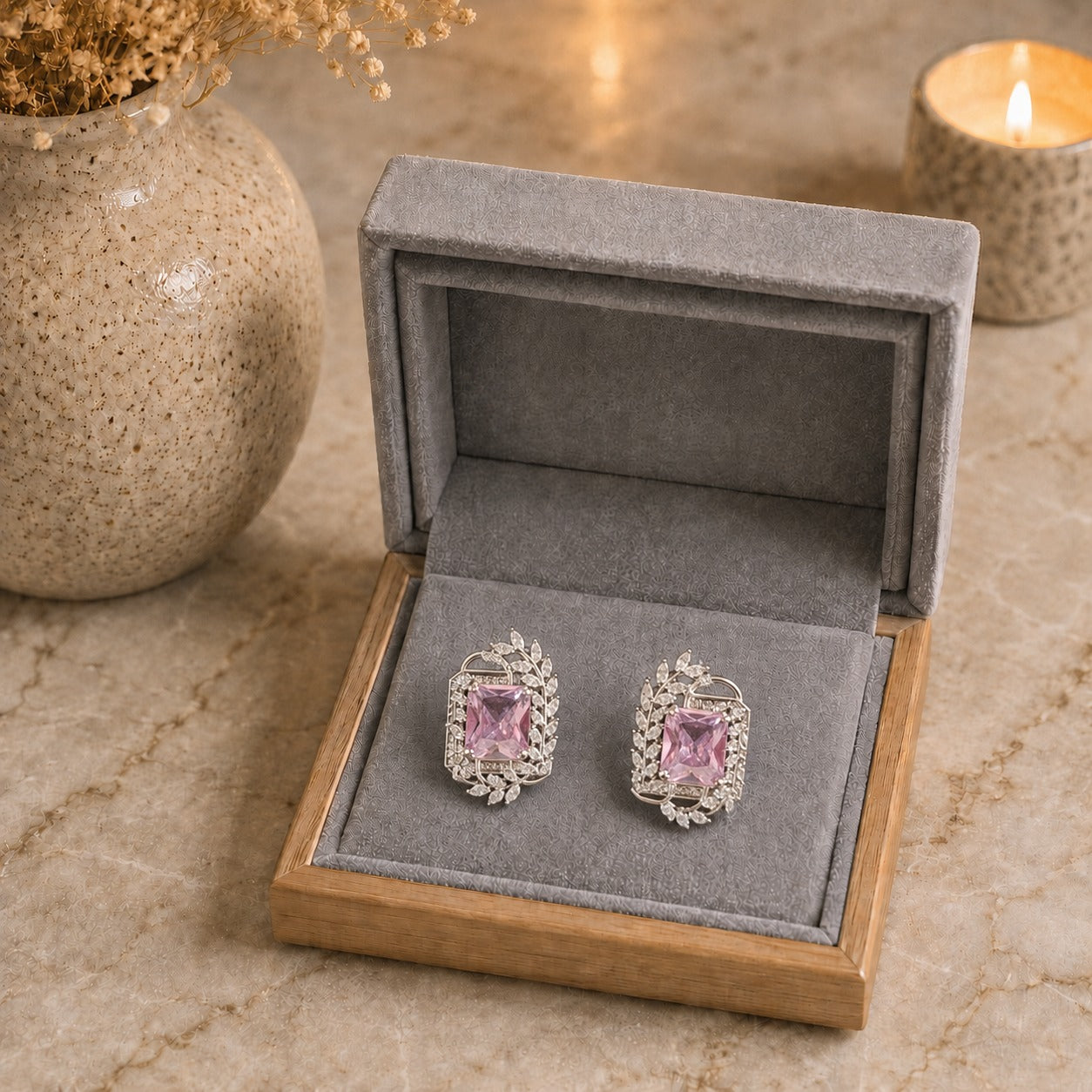 A pair of silver-toned stud earrings with rectangular pink gemstones and floral silver settings, displayed inside an open gray velvet jewelry box on a beige stone surface with a lit candle and vase in the background.