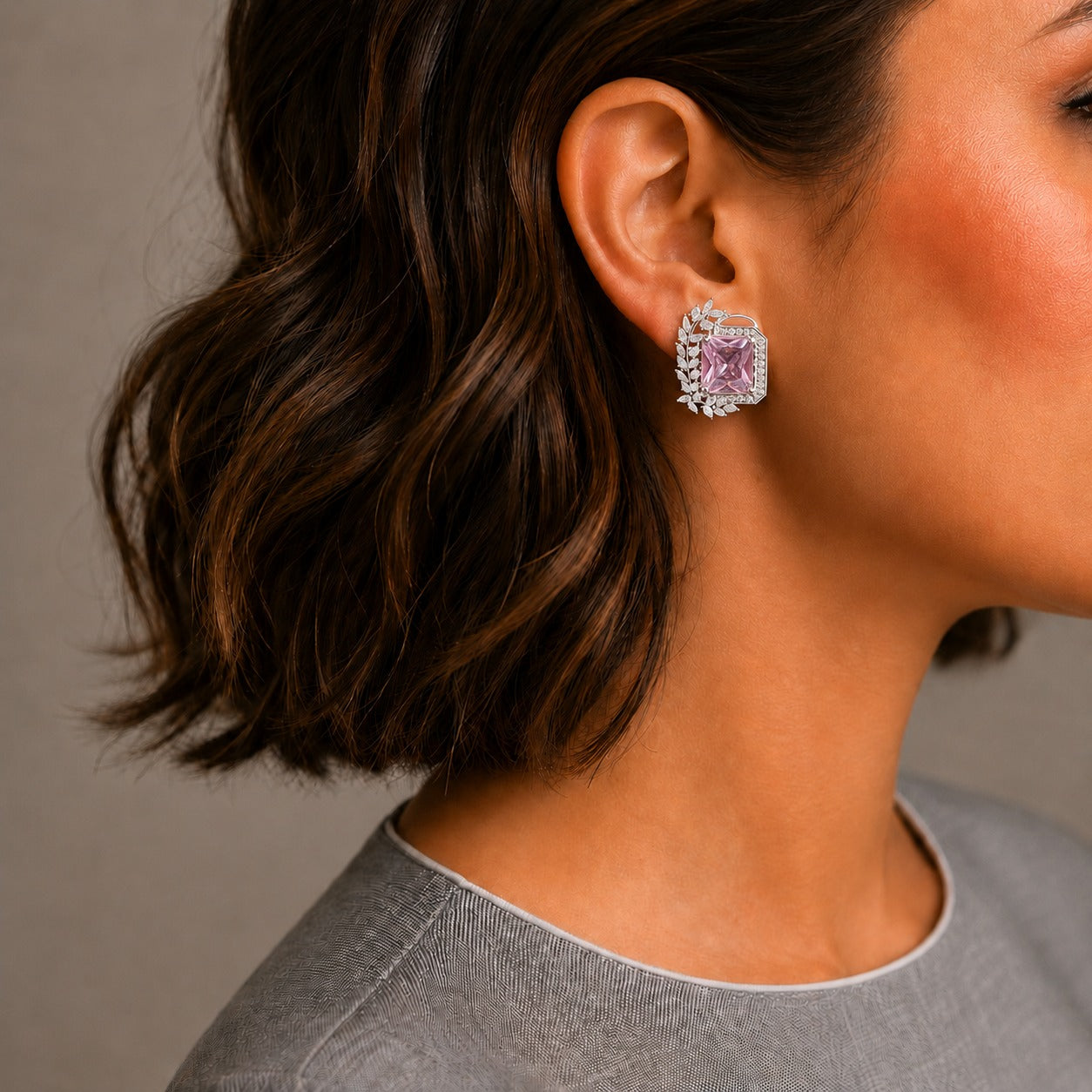 A close-up side profile of a woman with short wavy brown hair wearing a single square stud earring with a pink center stone and a halo of clear stones, against a neutral gray background.