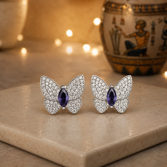 Silver butterfly stud earrings featuring pavé crystal wings and a violet marquise-cut centre stone displayed on a luxury stone surface