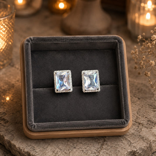 Emerald-cut crystal halo stud earrings displayed in a premium charcoal velvet jewelry tray on a natural stone surface with warm candlelight.