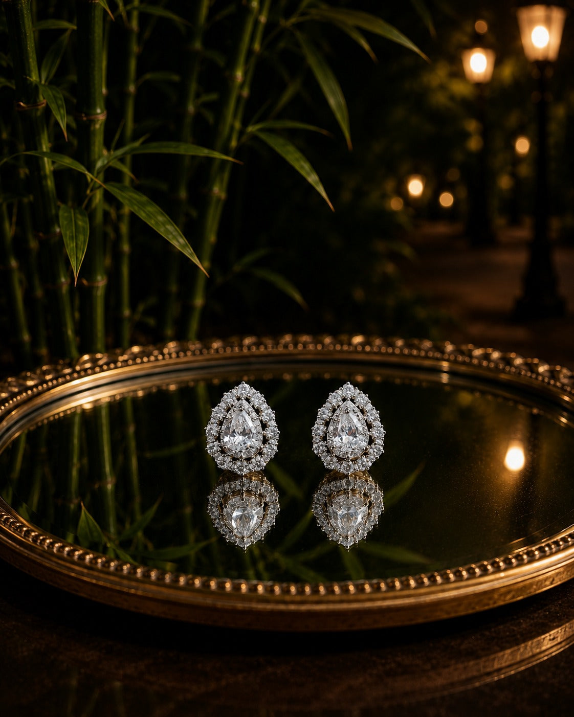 Pear-shaped double halo crystal stud earrings displayed on an antique mirror tray with bamboo, warm lantern lights, and a softly blurred evening garden background.