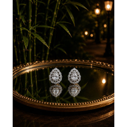 Pear-shaped double halo crystal stud earrings displayed on an antique mirror tray with bamboo, warm lantern lights, and a softly blurred evening garden background.