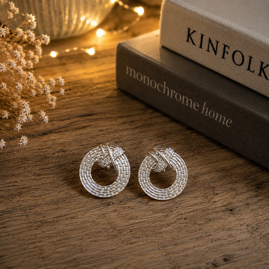 Pair of silver textured knot stud earrings displayed on a rustic wooden surface with decorative books and warm ambient lighting.