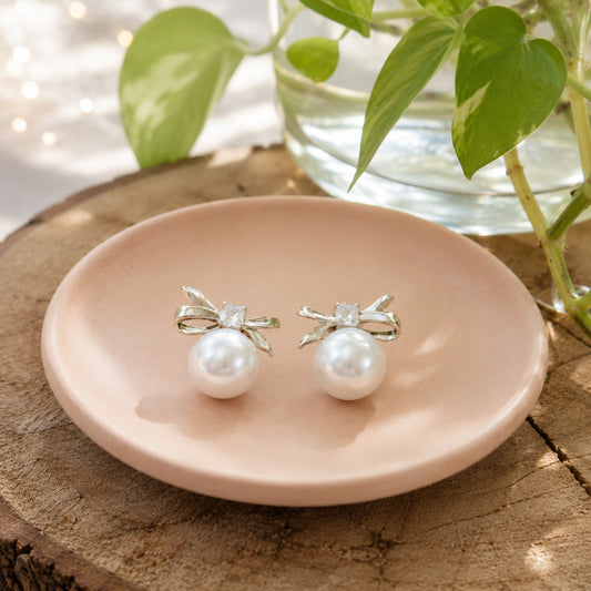 Silver bow pearl stud earrings with luminous pearl drops displayed on a blush ceramic plate in a bright natural setting.