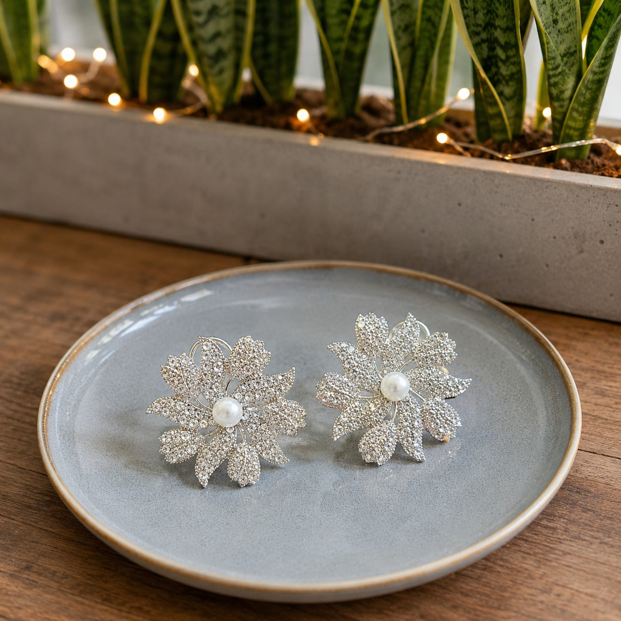 Pair of silver crystal floral pearl stud earrings with a pearl center displayed on a soft blue ceramic plate in a natural lifestyle setting.