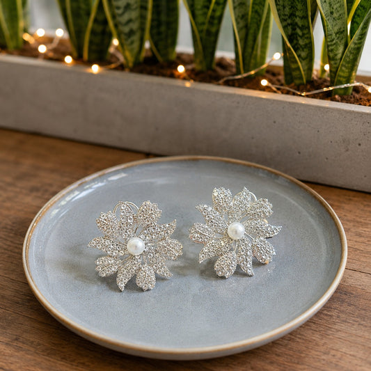 Pair of silver crystal floral pearl stud earrings with a pearl center displayed on a soft blue ceramic plate in a natural lifestyle setting.