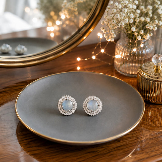 Round moonstone halo stud earrings displayed on a matte grey ceramic plate with a premium vanity background.