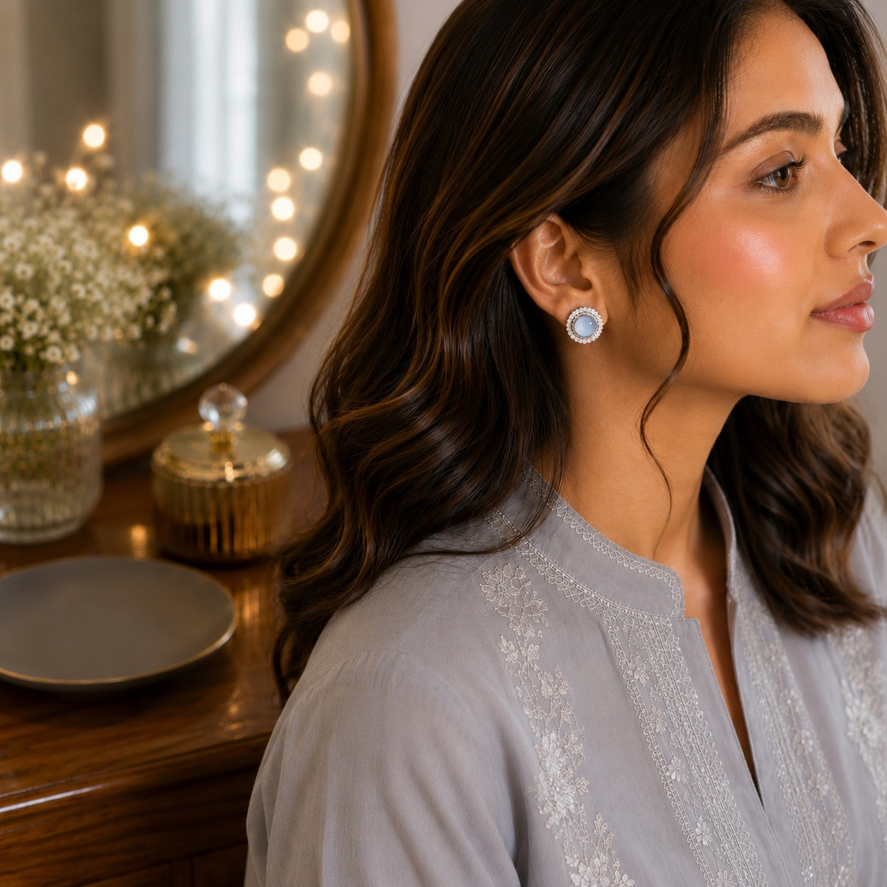 Woman wearing round moonstone halo stud earrings with a soft grey embroidered outfit in a luxury dressing table setting.