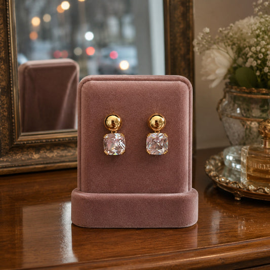 Gold-tone cushion crystal drop earrings displayed on a premium dusty rose velvet jewellery stand with a luxury dressing table background