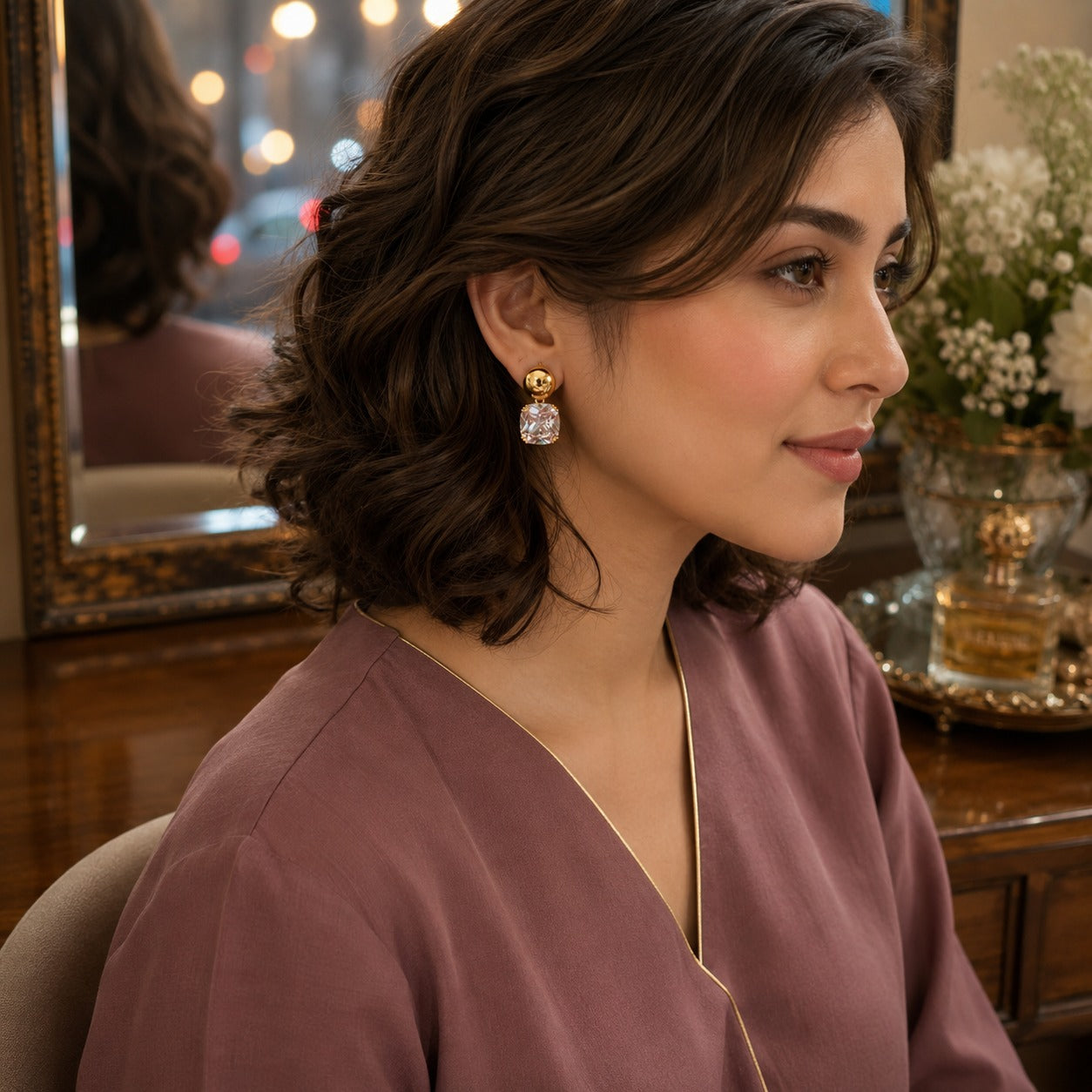 Woman wearing gold-tone cushion crystal drop earrings with a mauve V-neck outfit in a luxury indoor setting.