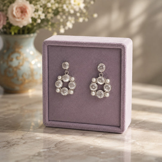 Silver crystal and pearl halo drop earrings displayed on a premium lavender velvet jewellery stand with a soft floral luxury background.