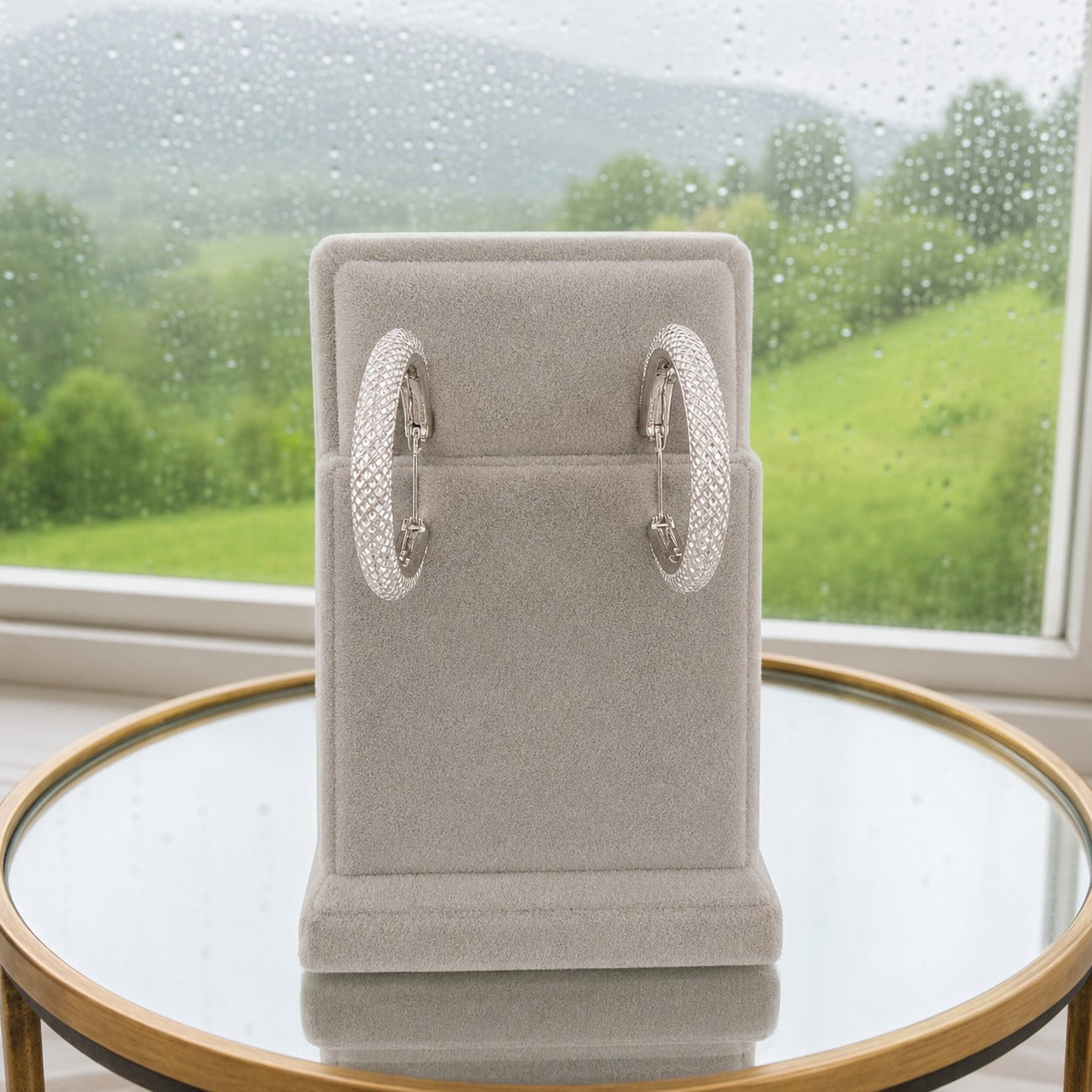 Silver textured crystal hoop earrings displayed on a premium light grey velvet stand with a rainy window backdrop.