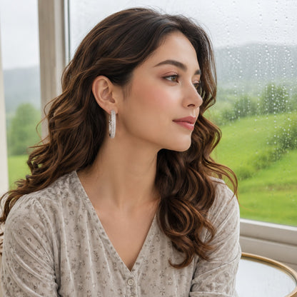 Woman wearing elegant silver textured crystal hoop earrings with soft waves and a light grey printed outfit beside a rain-covered window.