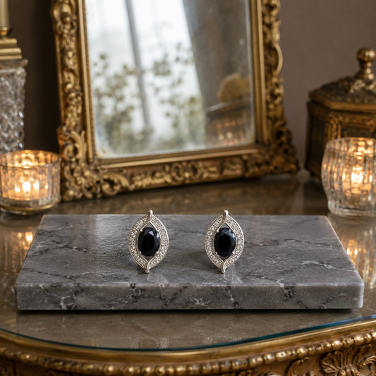 Black oval gemstone crystal halo latch-back earrings displayed on a polished grey marble slab with a warm antique mirror and candlelit luxury background.