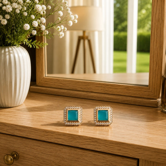 Premium square halo turquoise crystal stud earrings with princess-cut centre stone displayed on a polished wooden dressing table in a luxury lifestyle setting with floral décor and natural daylight.
