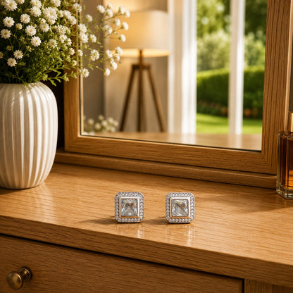 Premium square halo crystal stud earrings with princess-cut centre stone displayed on a polished wooden dressing table in a luxury lifestyle setting with floral décor and natural daylight.