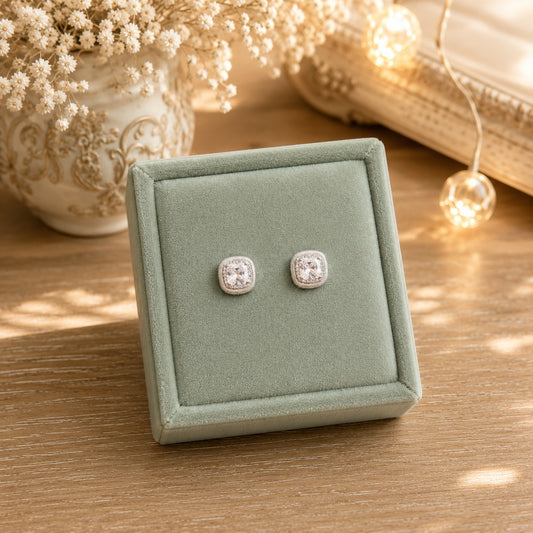 Silver cushion-cut halo zircon stud earrings displayed on a premium sage green velvet jewellery stand with warm luxury décor, fairy lights, and floral accents.