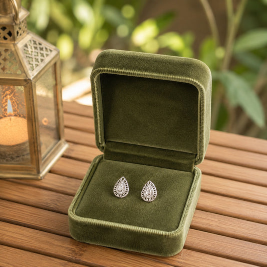 Silver teardrop crystal stud earrings displayed in an olive green velvet jewellery box on a wooden table with a lantern and blurred garden background.