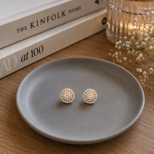 Round halo crystal stud earrings with gold-tone detailing displayed on a matte grey ceramic plate in a premium luxury jewelry setting.
