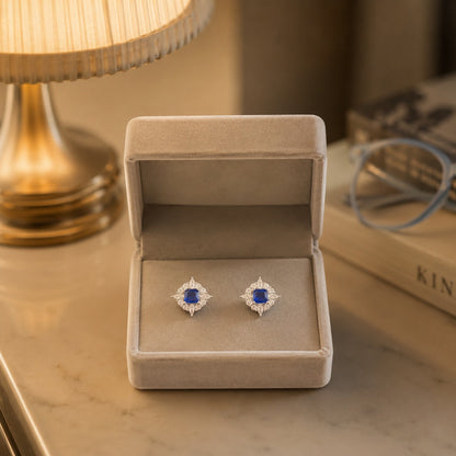 Crystal halo square gemstone stud earrings with a blue centre stone displayed in a premium beige velvet jewellery box under warm ambient lighting.