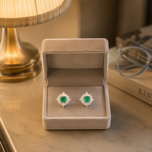 Crystal halo square gemstone stud earrings with an emerald green centre stone displayed in a premium beige velvet jewellery box under warm ambient lighting.