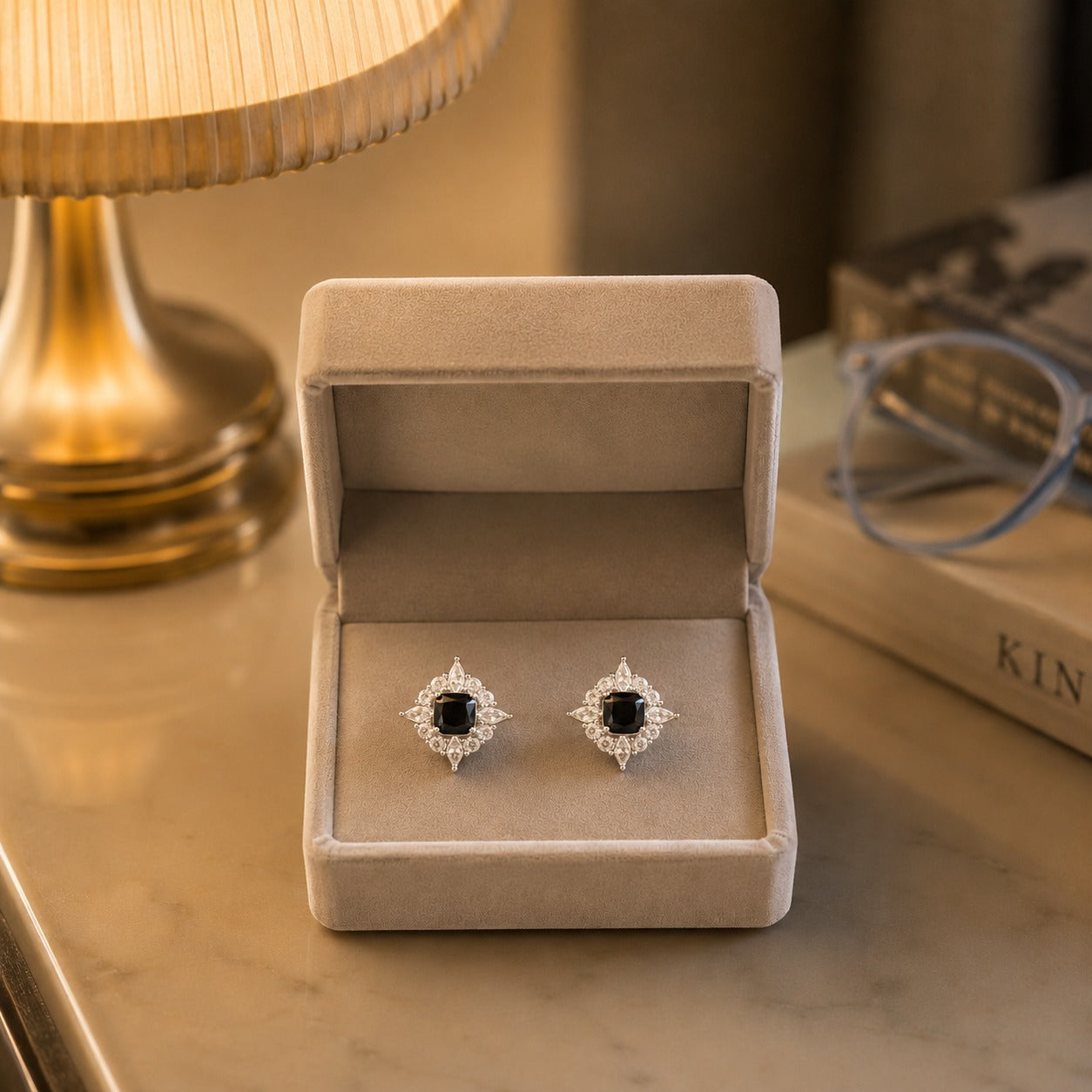 Crystal halo square gemstone stud earrings with a black centre stone displayed in a premium beige velvet jewellery box under warm ambient lighting.