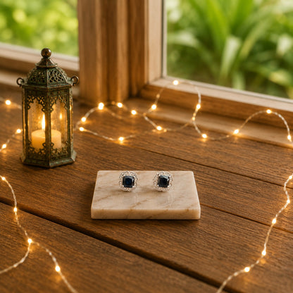 Halo cushion-cut crystal stud earrings with a midnight black centre stone displayed on a marble slab with warm decorative lighting and a luxury wooden background.