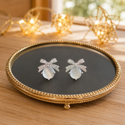 Silver bow stud earrings featuring natural Mother of Pearl cabochons displayed on a premium black mirrored jewellery tray.