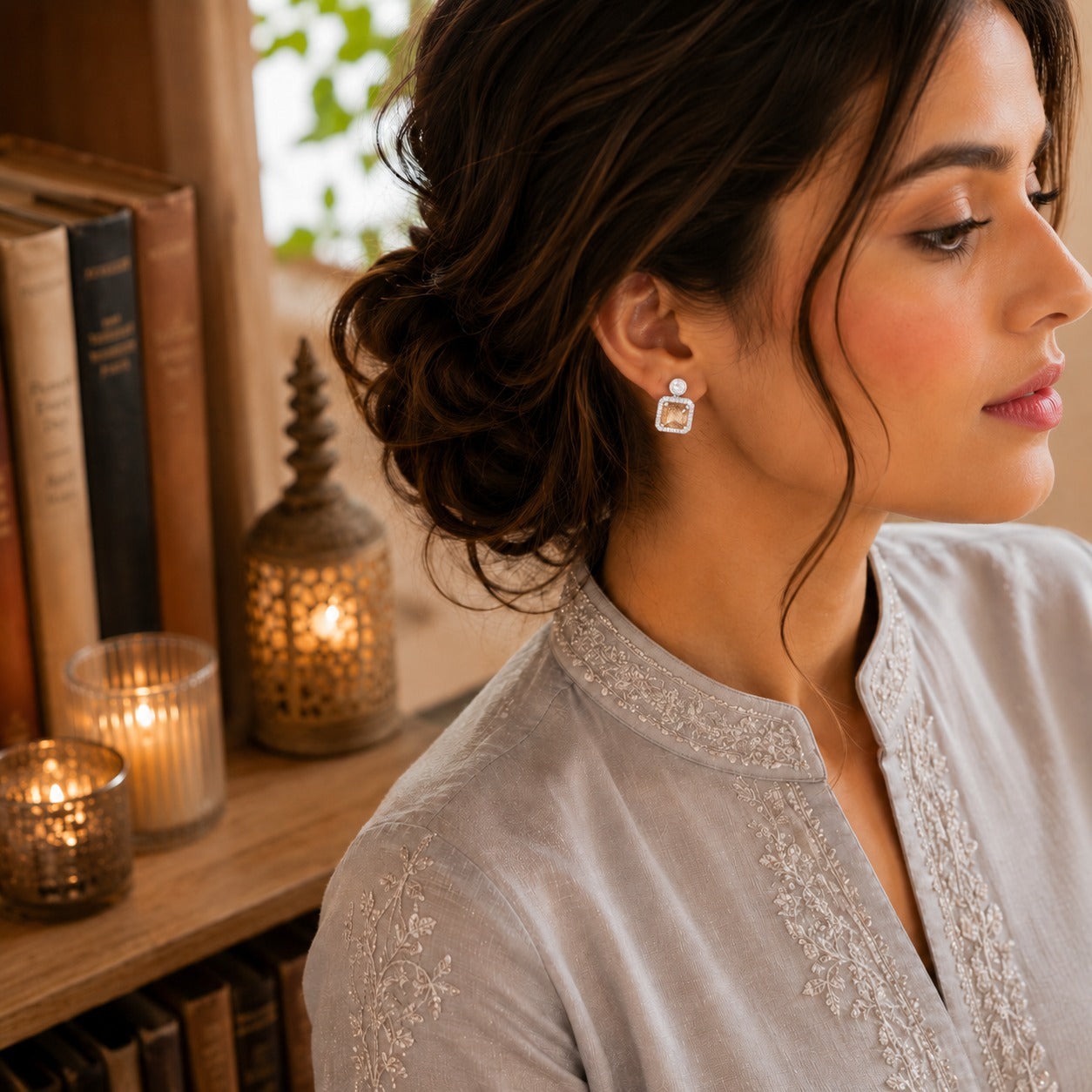 Woman wearing silver-tone square halo zircon stud earrings with a cushion-cut champagne centre stone and sparkling crystal border, styled with a traditional outfit in a warm luxury indoor setting.