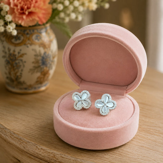 Silver-tone mother-of-pearl floral stud earrings displayed in a premium blush pink velvet jewelry box on a light oak table with a floral background.