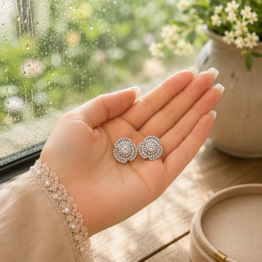 Silver crystal floral stud earrings displayed in a woman's hand beside a premium beige jewellery box with a softly blurred luxury window background