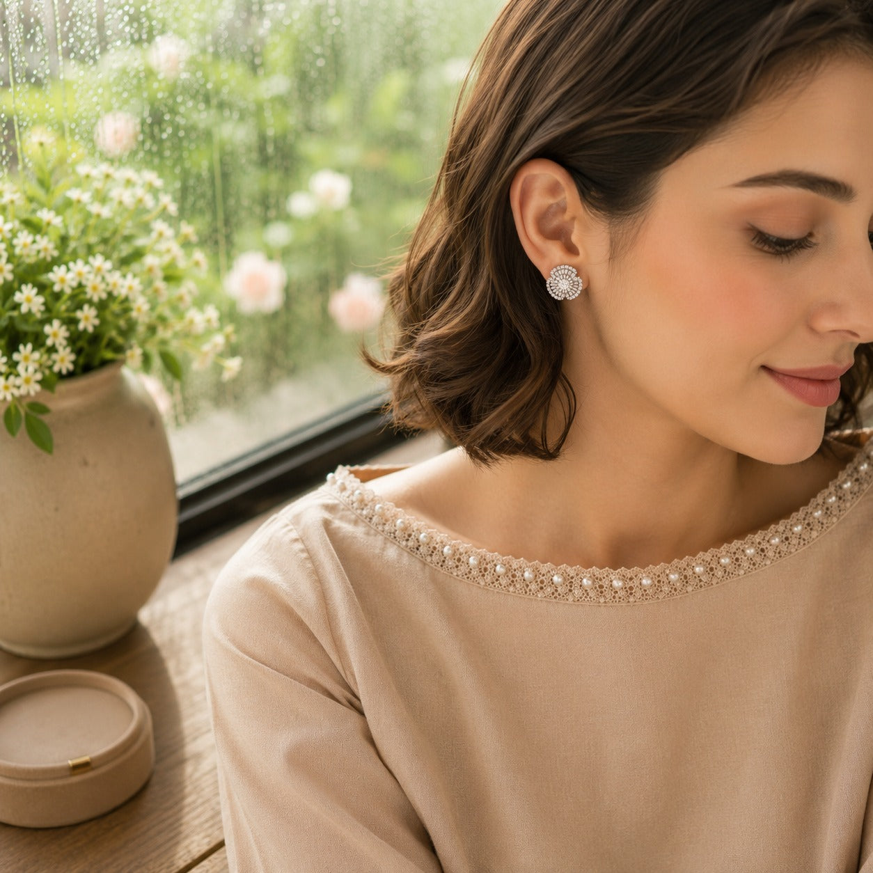 Woman wearing silver crystal floral stud earrings with a soft beige pearl-trim outfit in a luxury window-side lifestyle setting, showcasing sparkling floral earrings in a close-up side profile.
