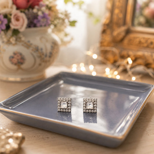Silver square halo crystal stud earrings with princess-cut clear centre stones displayed on a premium blue ceramic tray in a warm luxury setting