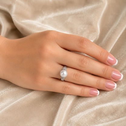 A close-up of a hand with French-tipped nails wearing a silver ring with a white pearl, resting on a draped beige velvet fabric.