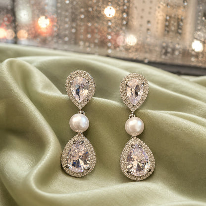 Silver statement earrings featuring double pear-cut crystals with pavé halos and luminous pearl connectors displayed on luxurious sage green satin fabric