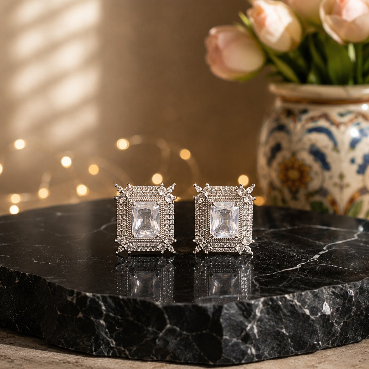 A pair of silver-toned square stud earrings with clear central stones and small clear stones around the edges, displayed on a black marble slab with a blurred floral vase and warm bokeh lights in the background.