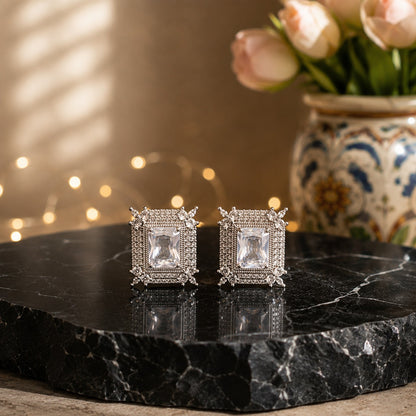 A pair of silver-toned square stud earrings with clear central stones and small clear stones around the edges, displayed on a black marble slab with a blurred floral vase and warm bokeh lights in the background.