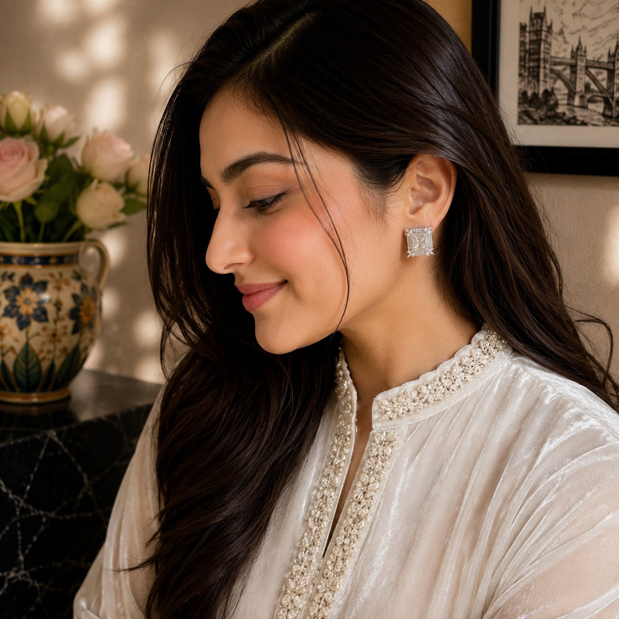 A woman with long dark hair is shown in profile wearing a silver-toned square stud earring with a clear central stone, paired with a cream embroidered top. The background features warm sunlight, a floral vase, and framed wall art.