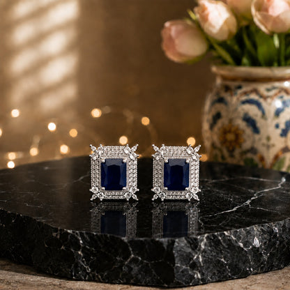 A pair of rectangular stud earrings with deep blue center stones and silver-toned pavé borders, displayed on a black marble slab with a blurred floral vase in the background.