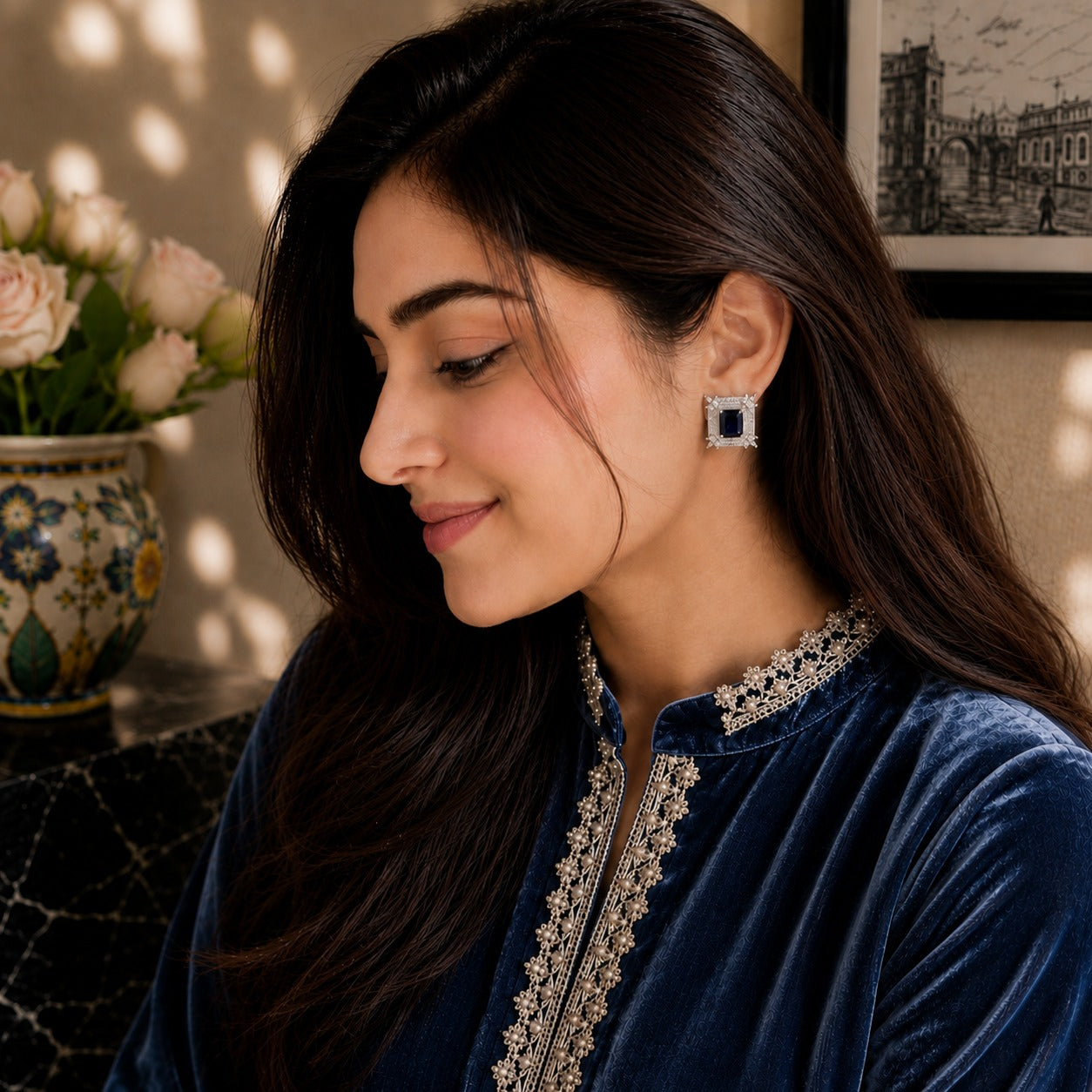 A woman in profile wearing a navy blue velvet top with ornate silver embroidery and a matching silver square stud earring with a dark blue center, set against a warm indoor background with a vase of flowers.