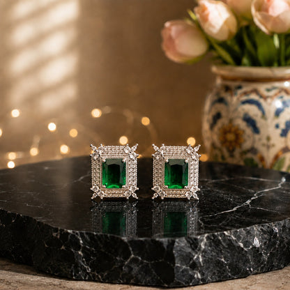 A pair of square stud earrings with emerald-cut green gemstones surrounded by small clear stones in a silver-toned setting, displayed on a black marble slab with a blurred floral vase in the background.