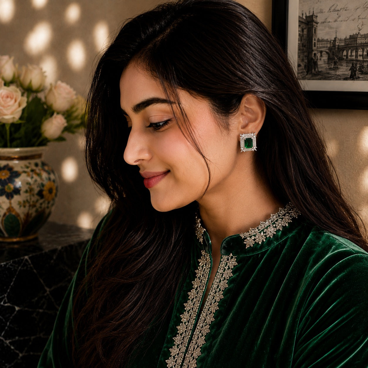 A woman in a dark green embroidered traditional outfit wearing a square green and silver earring, shown in a warm, sunlit indoor setting with a vase of flowers in the background.