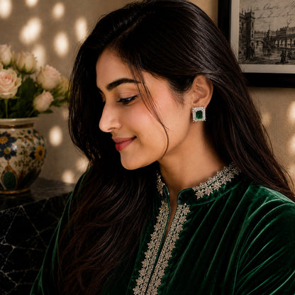 A woman in a dark green embroidered traditional outfit wearing a square green and silver earring, shown in a warm, sunlit indoor setting with a vase of flowers in the background.