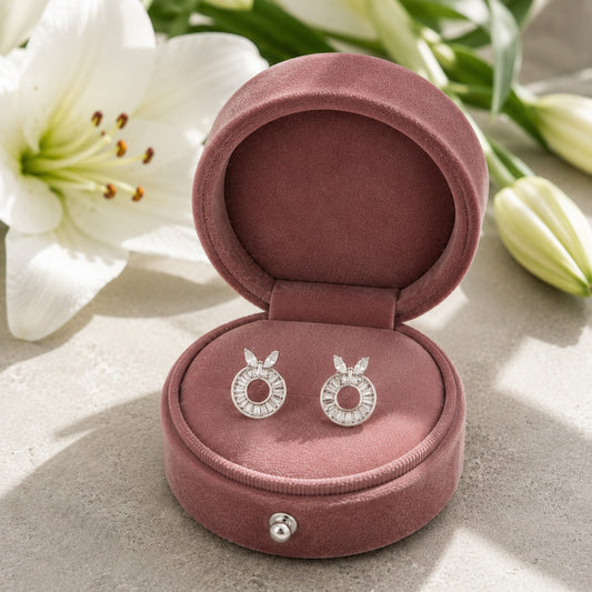 A pair of silver-toned hoop earrings with clear stones, displayed inside an open dusty rose velvet jewelry box. White lilies are softly blurred in the background on a light stone surface.