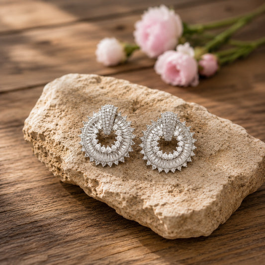 Silver circular statement stud earrings with baguette and round crystal detailing displayed on a natural stone surface.