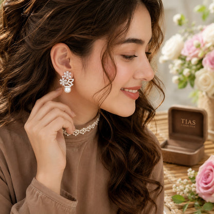 Model wearing silver floral pearl drop earrings with crystal blossoms and a pearl accent in a soft side-profile portrait.