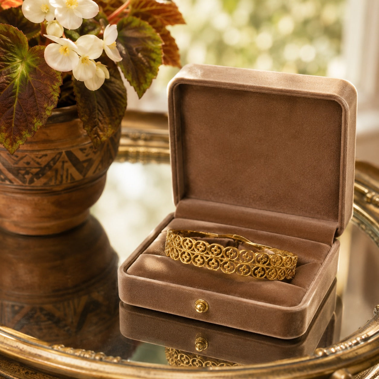 Gold openwork bracelet with a front magnetic clasp displayed inside a premium mocha velvet jewellery box on a mirrored tabletop beside fresh white flowers