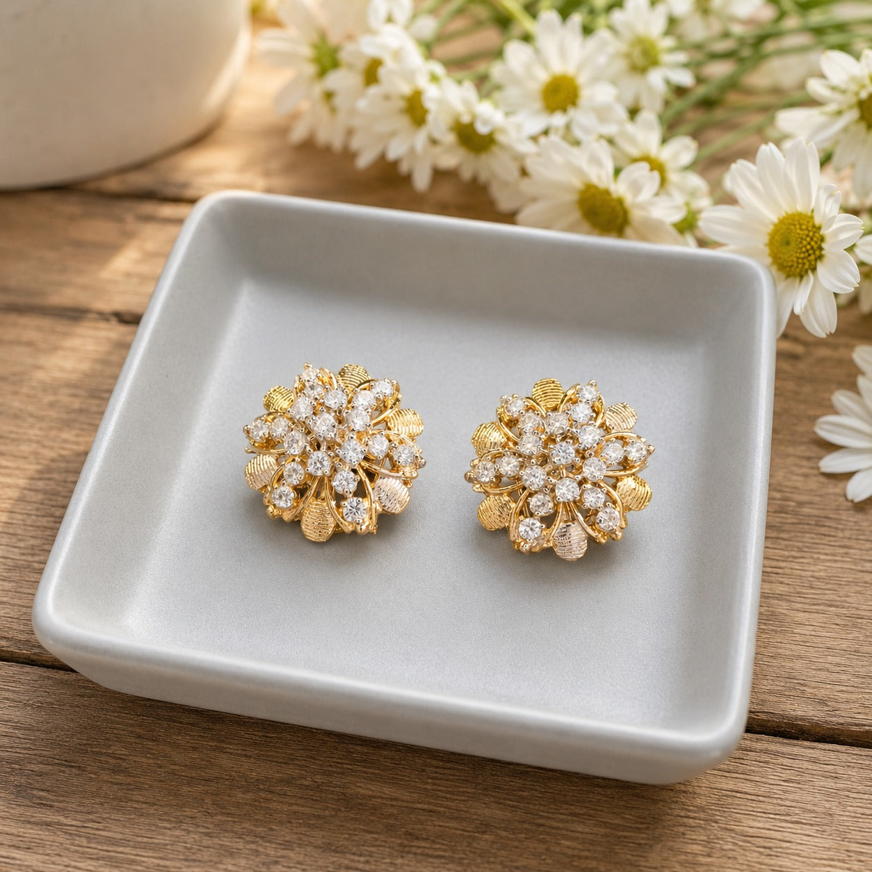 Gold floral crystal stud earrings with textured leaf detailing displayed on a white ceramic jewellery tray.